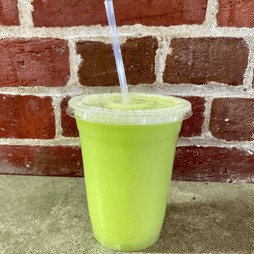 A vibrant green smoothie in a clear plastic cup with a straw, against a brick wall.