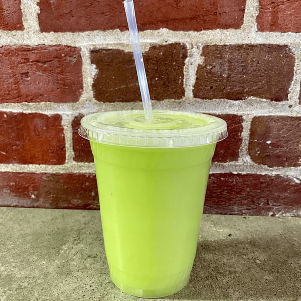 A vibrant green juice in a clear plastic cup with a straw, against a red brick wall.
